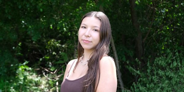 Young woman with long hair stands outdoors in a green, wooded area.