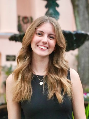 Young woman with long wavy hair smiling outdoors in front of a fountain.
