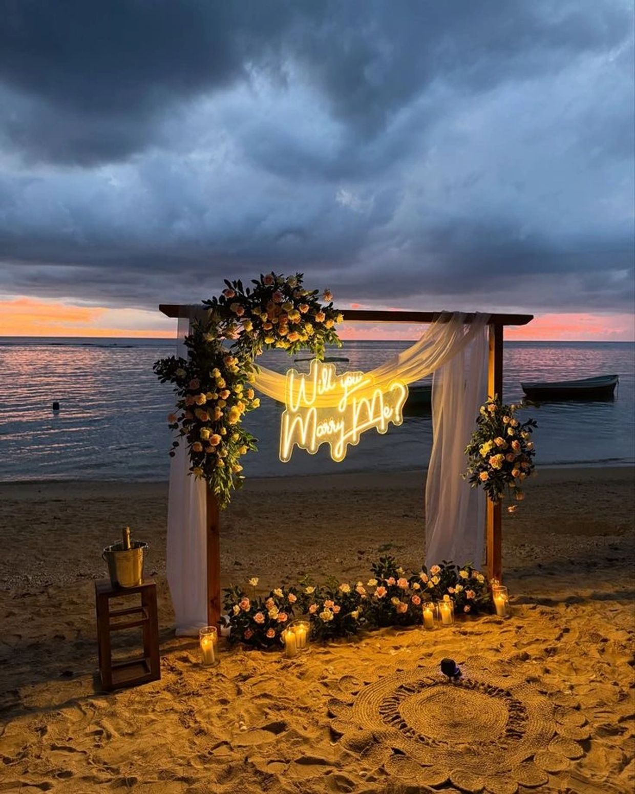 Romantic beach proposal setup with flowers and glowing "Will you marry me?" sign.