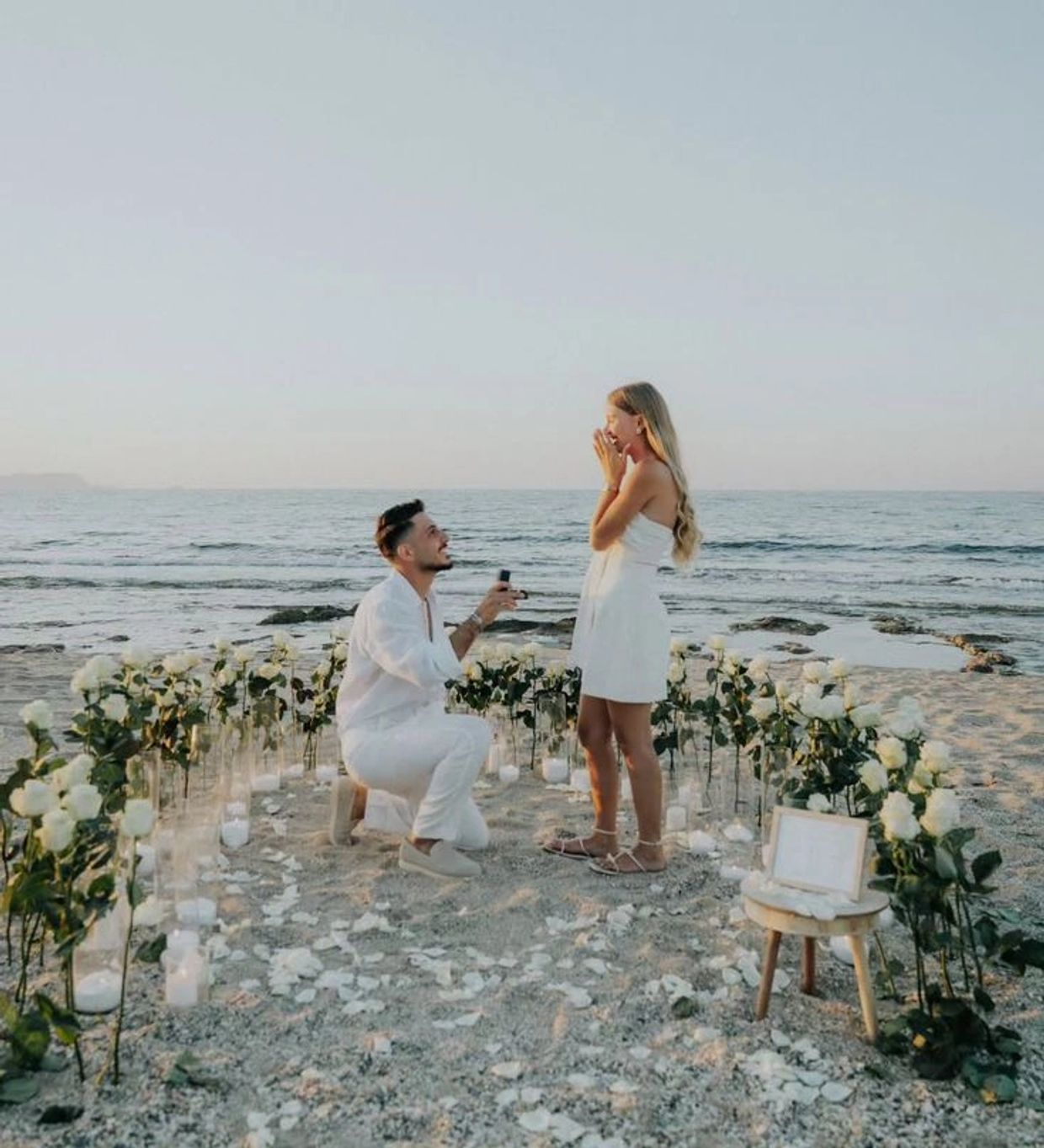 Man proposing to woman on a decorated beach with white roses and candles.