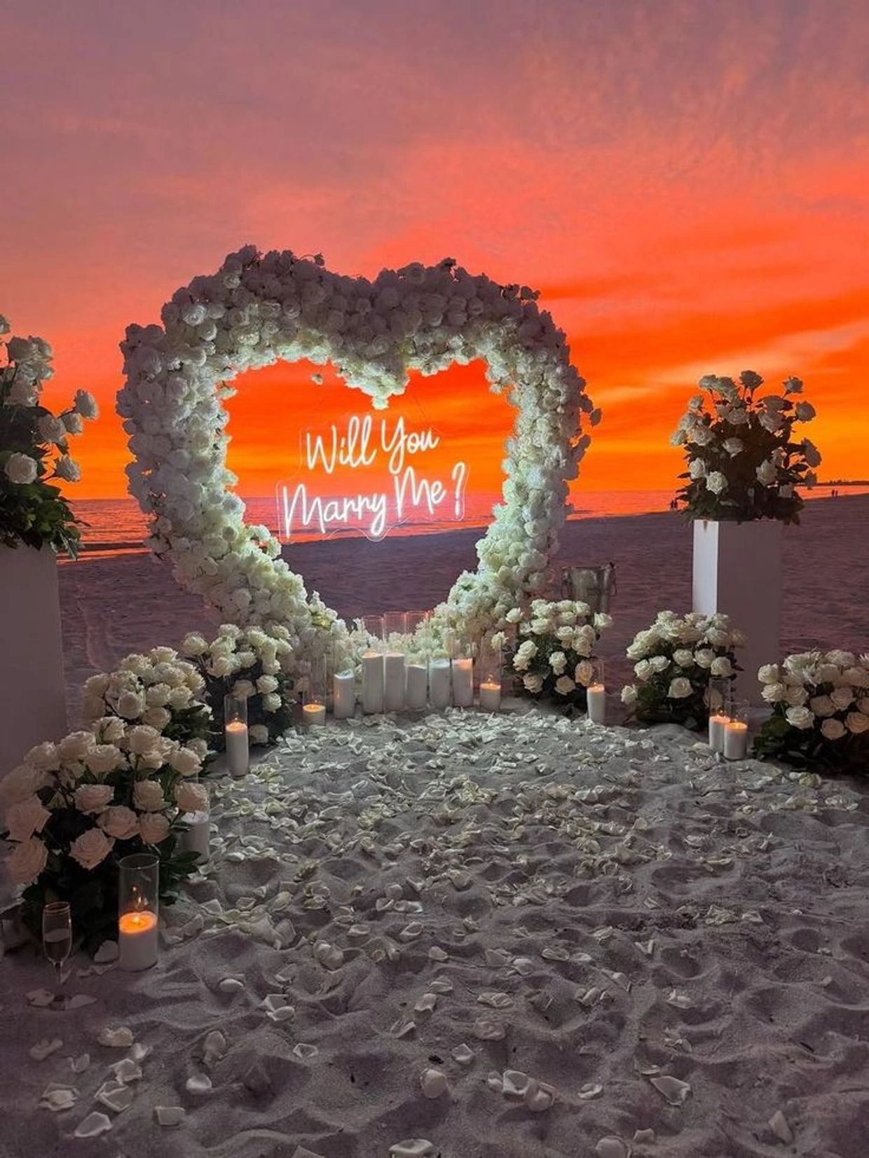 Romantic beach proposal setup with white roses and candles at sunset.