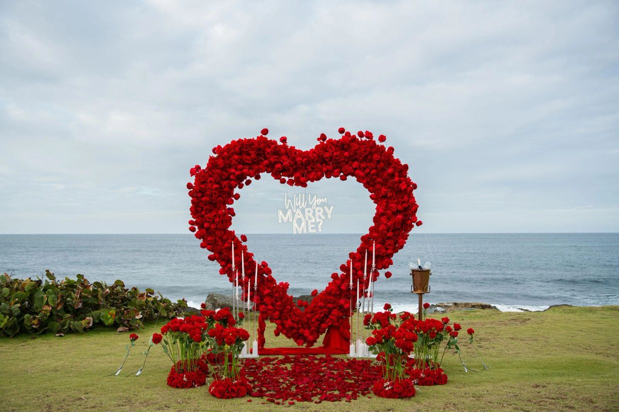 Heart-shaped rose arrangement by the sea with 'Will You Marry Me?' sign.