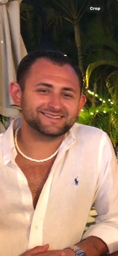 Smiling man in white shirt and shell necklace at a tropical outdoor setting.