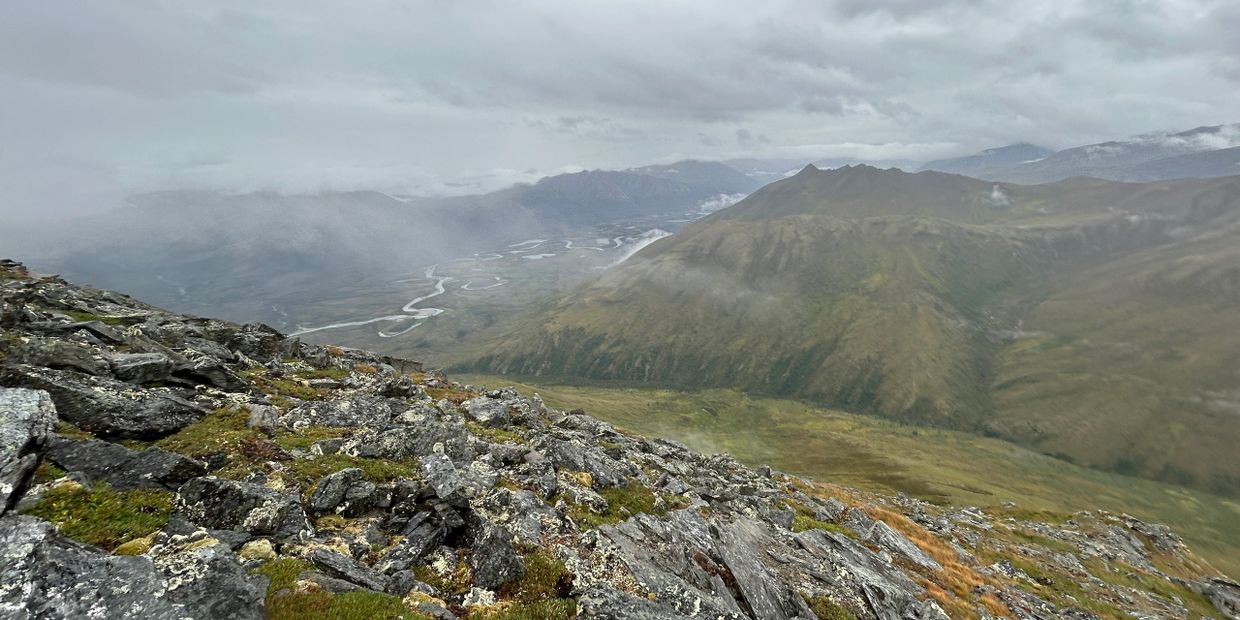 Rocky mountain slope overlooking a winding river valley under cloudy skies.