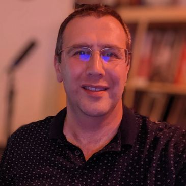 Smiling man wearing glasses and a dark polo shirt indoors.