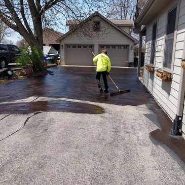 Worker sealing a driveway with a broom on a sunny day.