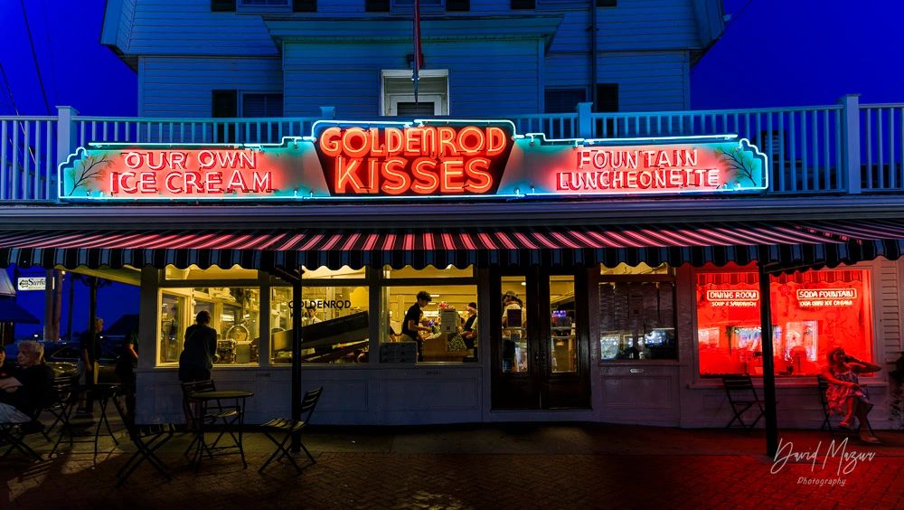 Neon-lit Goldenrod Kisses ice cream and luncheonette at night with people inside and outside.