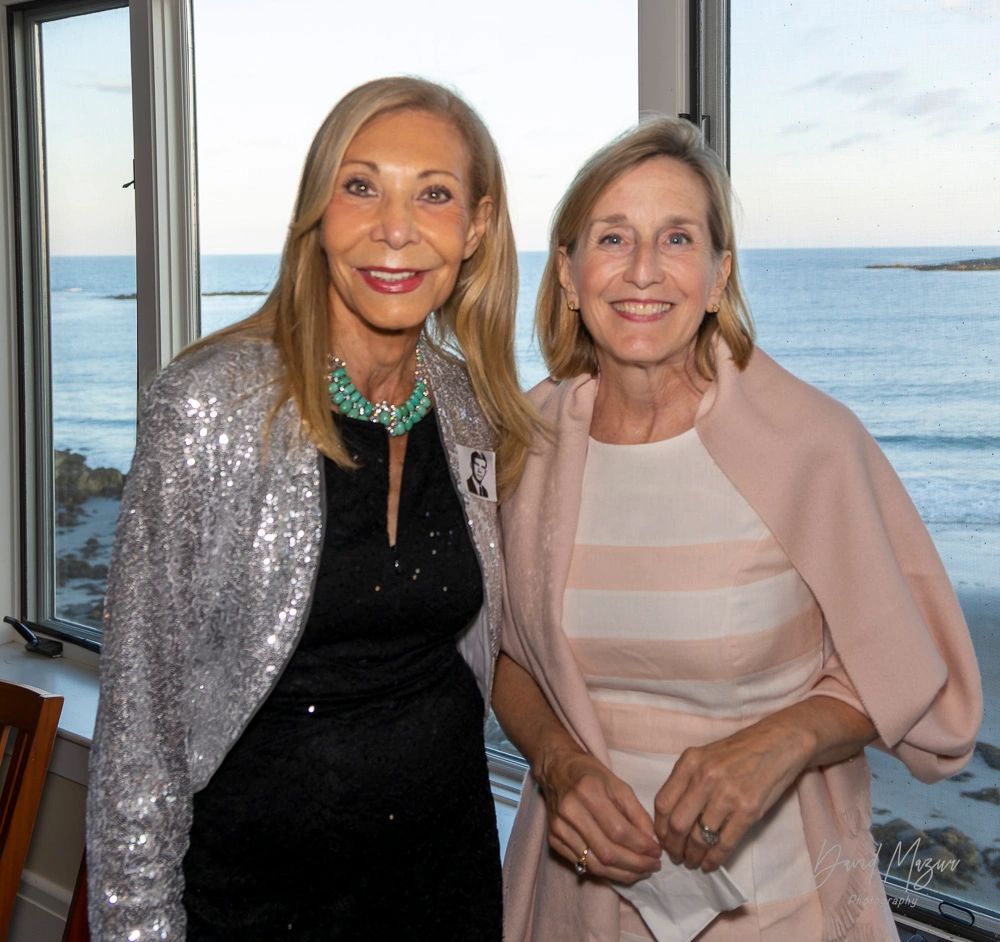 Two smiling women posing indoors with an ocean view in the background.