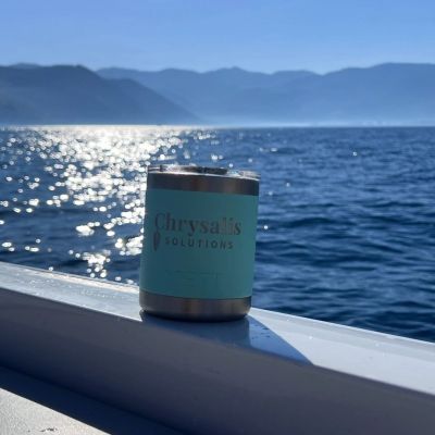 A stainless steel tumbler with 'Chrysalis Solutions' by a sparkling lake and mountains.
