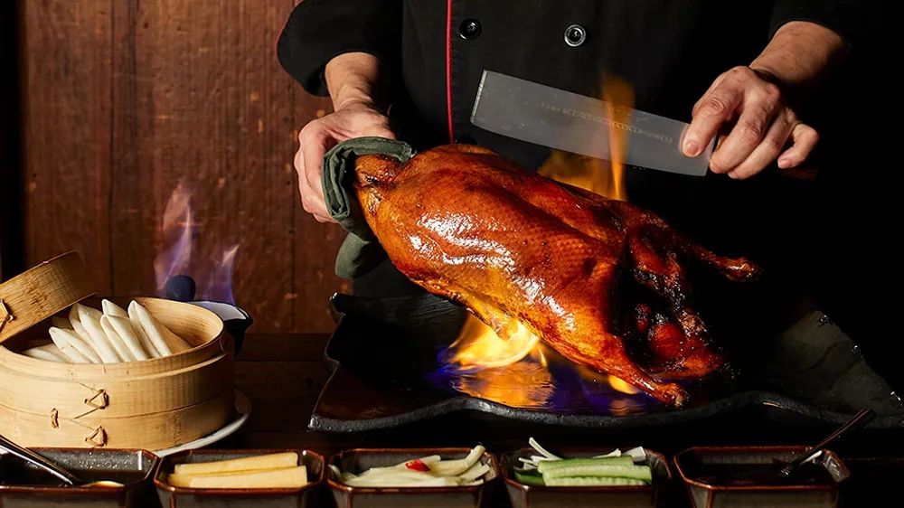 Chef holding a flaming roast duck with condiments and buns.
