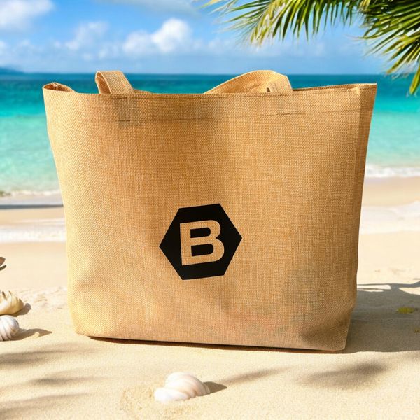 Beach tote bag with a bold 'B' logo on sandy shore under palm leaves.
