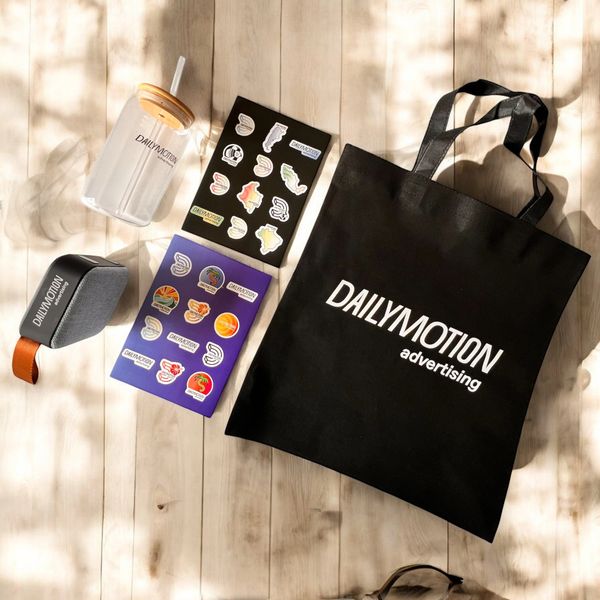 Branded Dailymotion merchandise including a tote bag, tumbler, speaker, and sticker sheets.