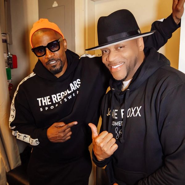 Two men in hoodies posing together, one wearing a bright orange beanie and sunglasses.