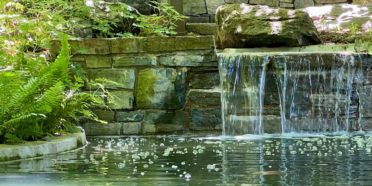 Small waterfall cascading over a stone wall into a pond surrounded by green plants.