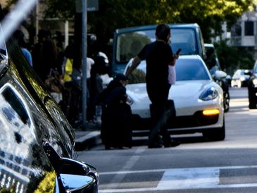 A person walking near a white car on a busy street with reflections on nearby vehicles.