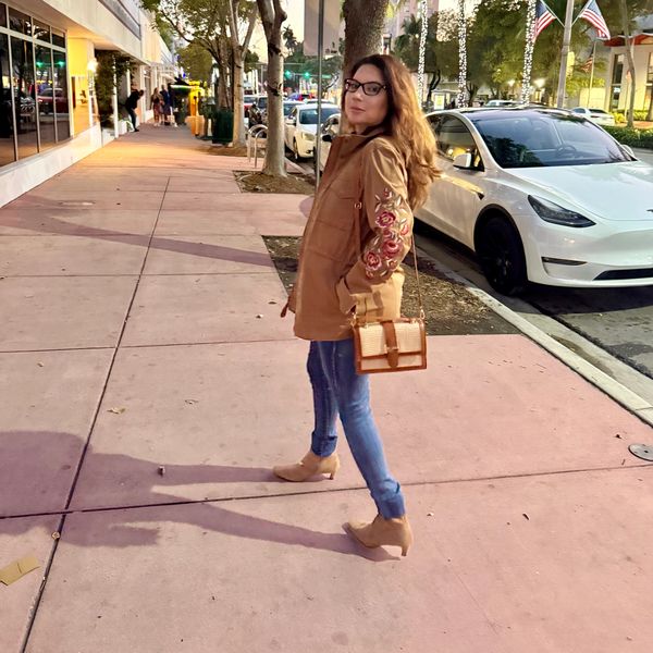 Woman in brown jacket and jeans walking on a city sidewalk at dusk.