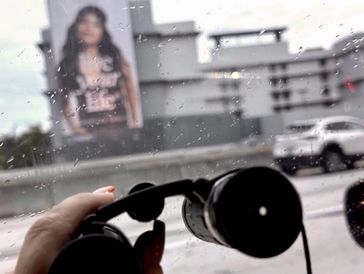 Hand holding binoculars looking through a rainy car window at a billboard.