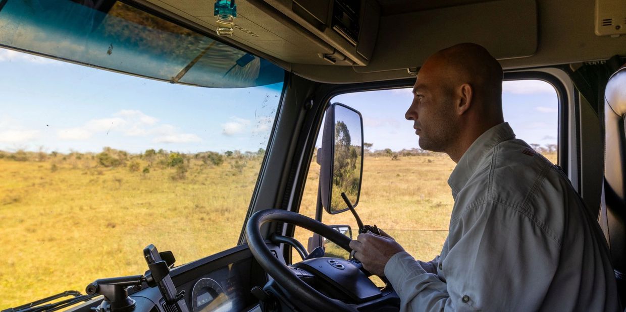 A man attentively drives a truck through a grassy landscape under a clear sky.