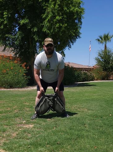 Man in a t-shirt and cap lifting heavy bags outdoors on grass.