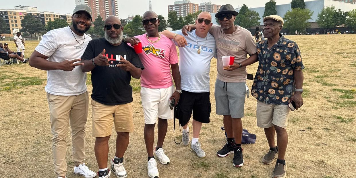 Six men casually posing together outdoors on a dry grassy field.