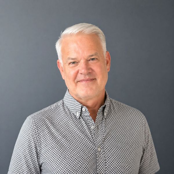 Smiling older man with white hair in a patterned shirt against a gray background.