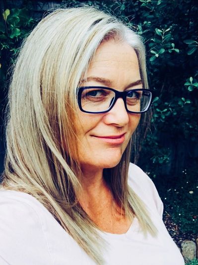 A woman with long blonde hair and black glasses smiling outdoors.