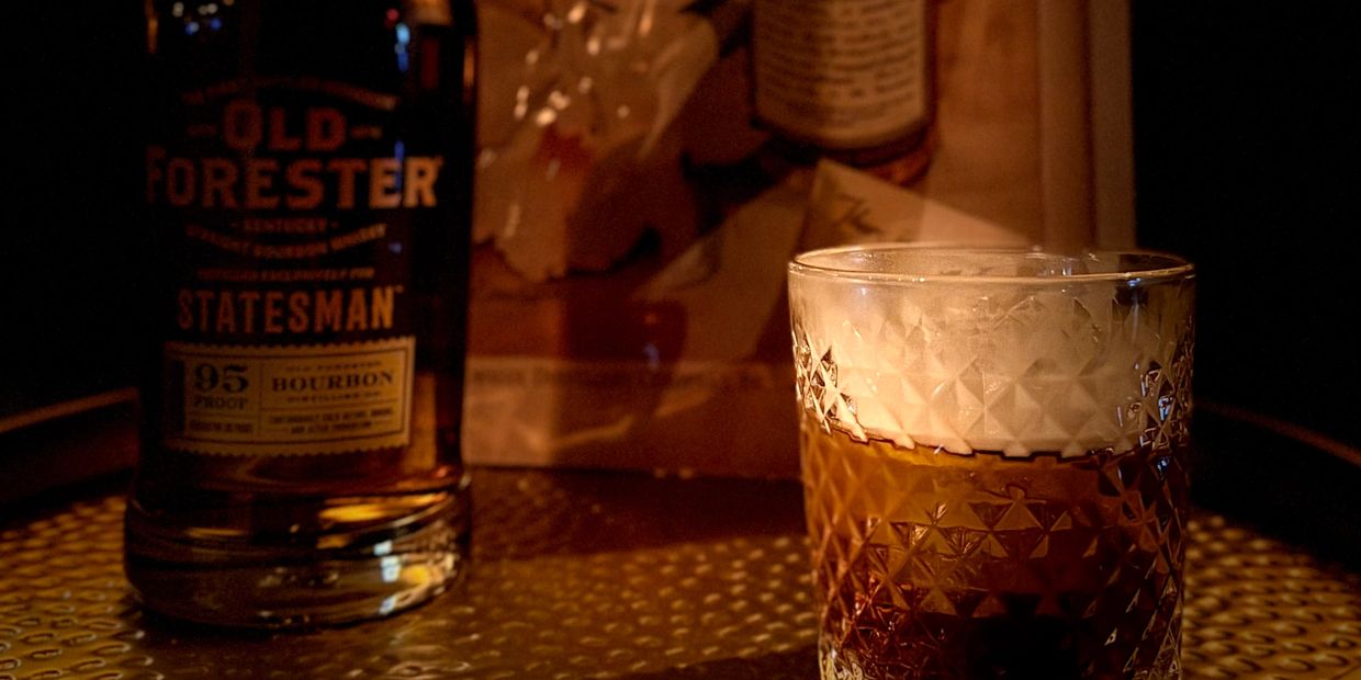A glass of bourbon whiskey with a bottle of Old Forester Statesman in warm lighting.