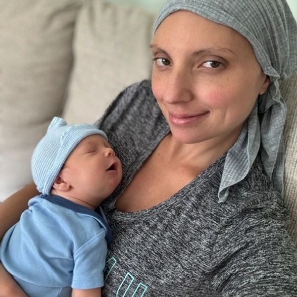 A smiling woman holds a sleeping newborn baby wearing a blue outfit and hat.