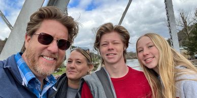 A happy family selfie outdoors on a bridge with a river and trees in the background.