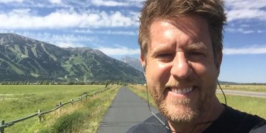 A man smiling outdoors on a sunny day with mountains in the background.