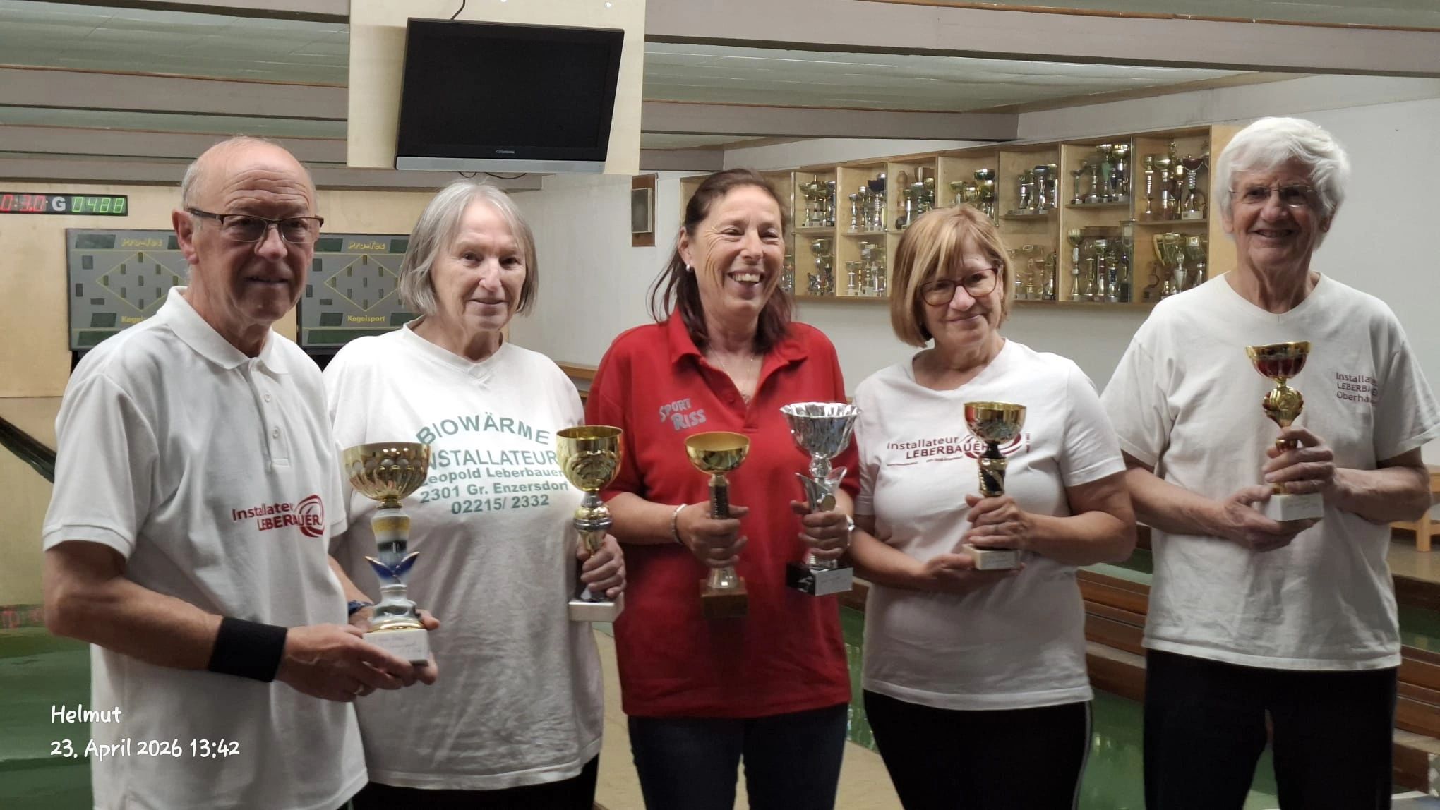 Five people proudly holding trophies in a bowling alley.