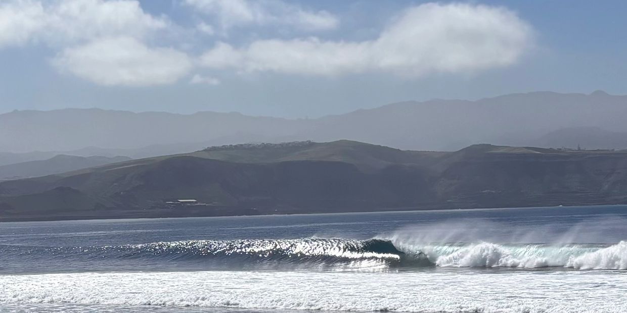 Shimmering ocean waves break near a mountainous coastline under a partly cloudy sky.