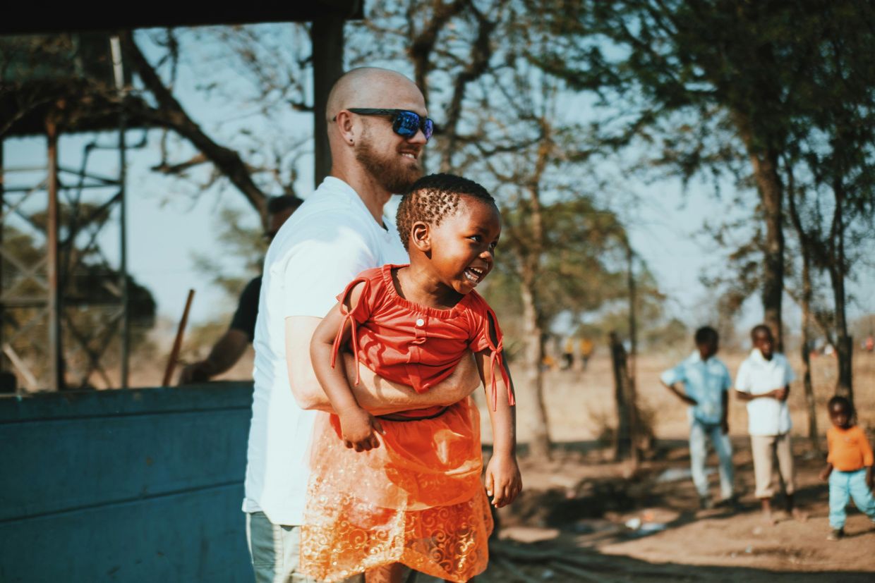 A man in sunglasses joyfully holds a laughing child in an outdoor setting.