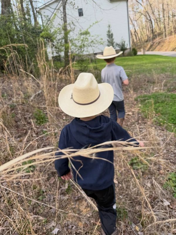 Two children wearing cowboy hats walk through dry grass near a house.