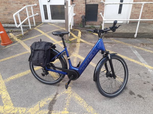 Blue Trek electric bike parked with a black pannier bag on a marked pavement.