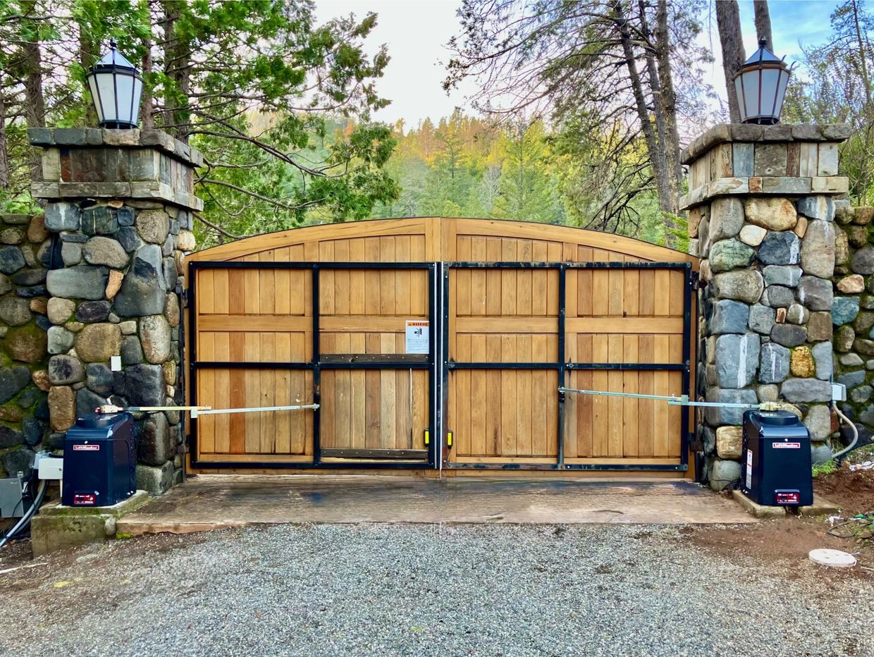Wooden automatic gate with stone pillars and lanterns, set in a forested area.