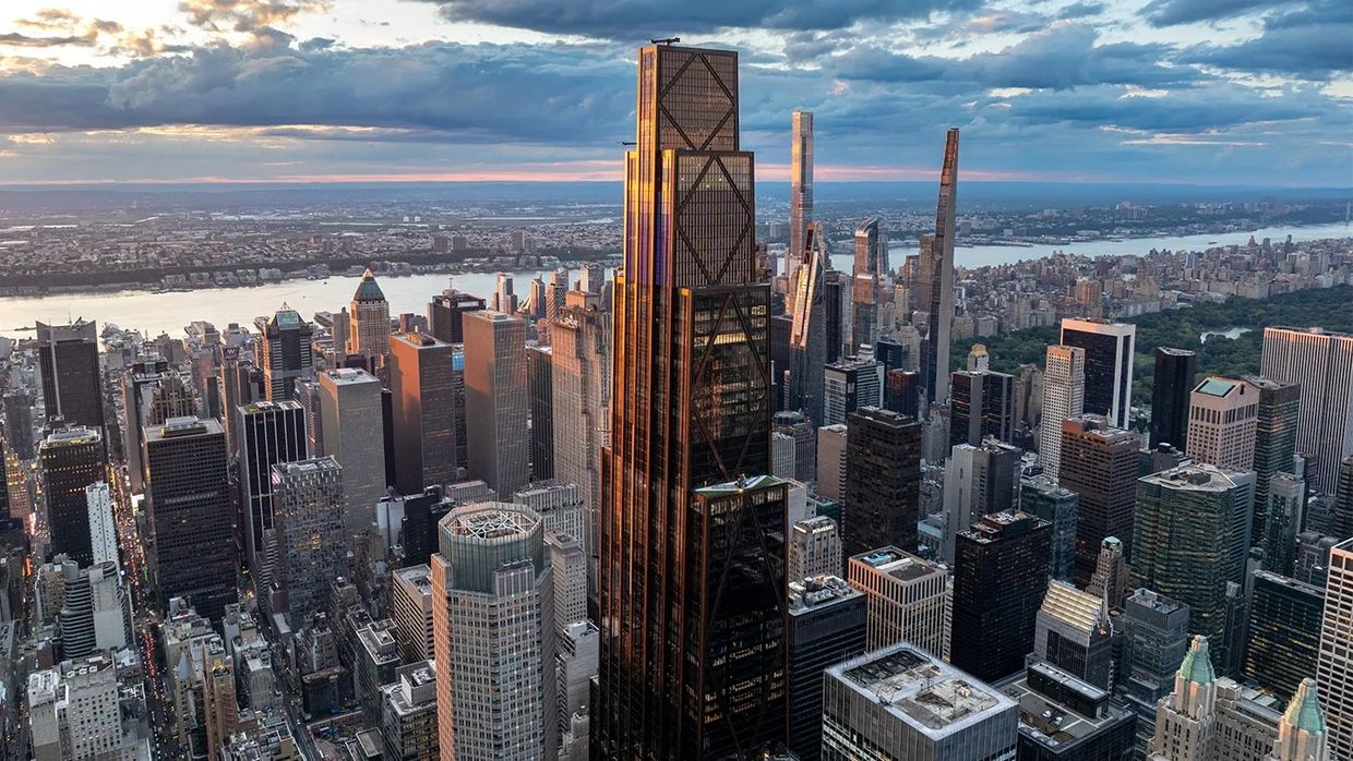 Skyline of a city with a tall, sunlit skyscraper and river in the background.