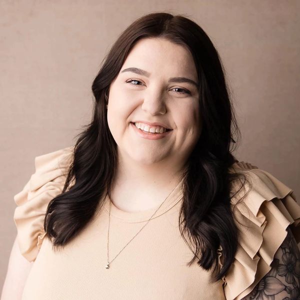 Smiling woman with dark hair and floral tattoo on arm in beige top.