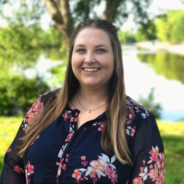 Smiling woman in a floral blouse outdoors near a lake.