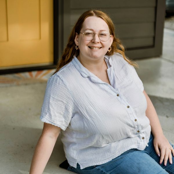 Smiling woman with glasses sitting outdoors near a yellow door.