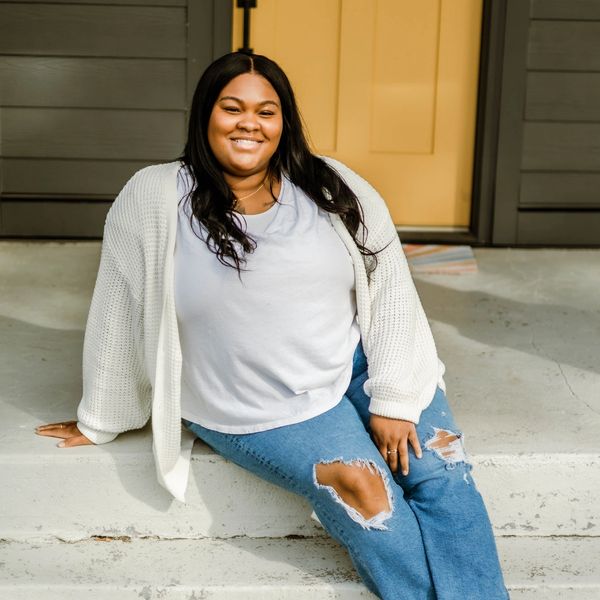 A smiling woman sits on outdoor steps wearing a white sweater and ripped jeans.