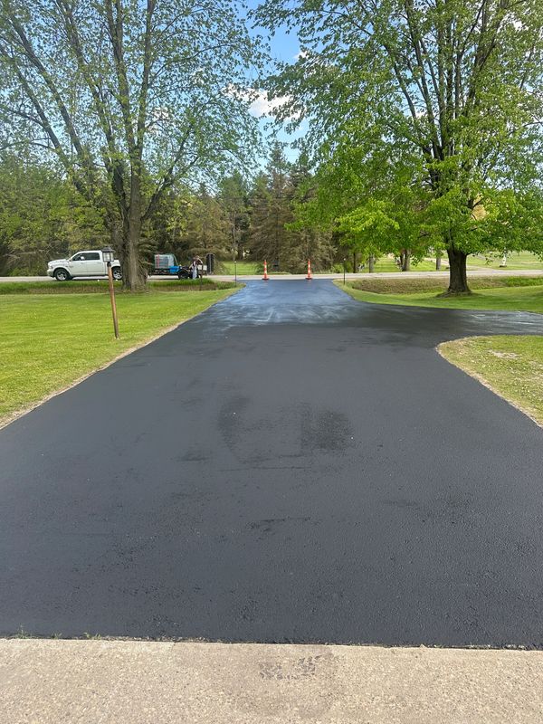 Freshly paved black driveway leading to a quiet road with trees on both sides.