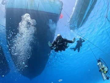 Two scuba divers ascend near a large boat underwater with air bubbles rising.