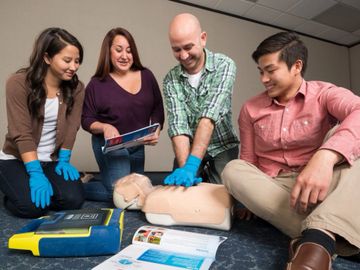 Group learning CPR on a mannequin with an AED trainer in a classroom.
