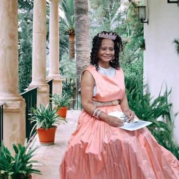 A woman in a peach dress and tiara sitting on a porch surrounded by plants.