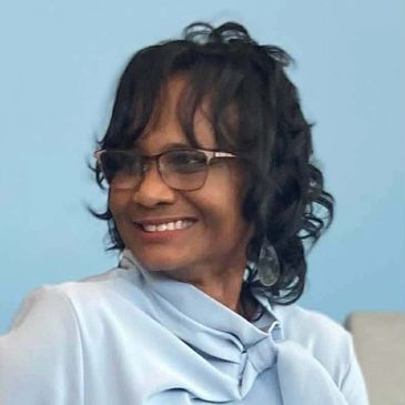 Smiling woman with glasses and curly hair in light blue blouse.