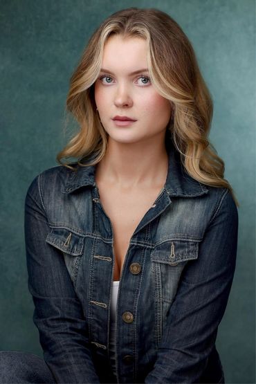 Young woman with blonde hair wearing a denim jacket against a teal background.