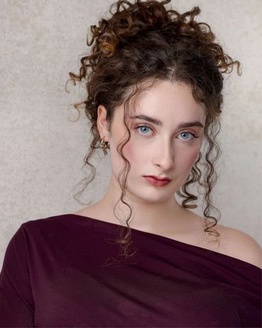 Portrait of a woman with curly hair and blue eyes in a purple off-shoulder top.