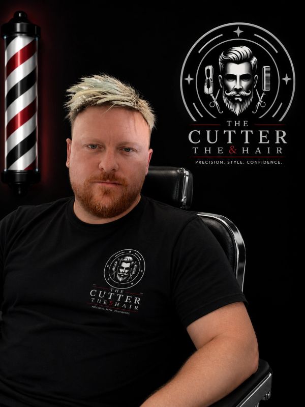 Man with styled hair and beard in barber chair with logo behind.