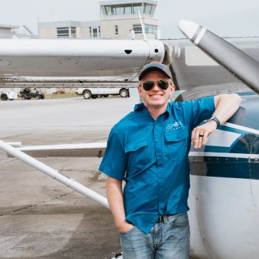 Smiling pilot in blue shirt stands beside small airplane on runway.